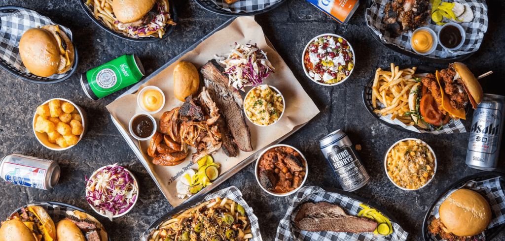 A flat lay of assorted BBQ dishes including brisket, ribs, burgers, fries, slaw, and corn. Beverages like canned soda and beer are scattered around. The food is served on trays and in baskets with checkered paper on a dark, textured surface.