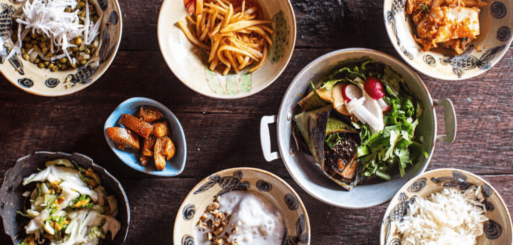 A wooden table set with various Asian dishes, including leafy salad, kimchi, pickled vegetables, shredded coconut, sliced fruit, and rice. The bowls and plates are decorated with intricate patterns.