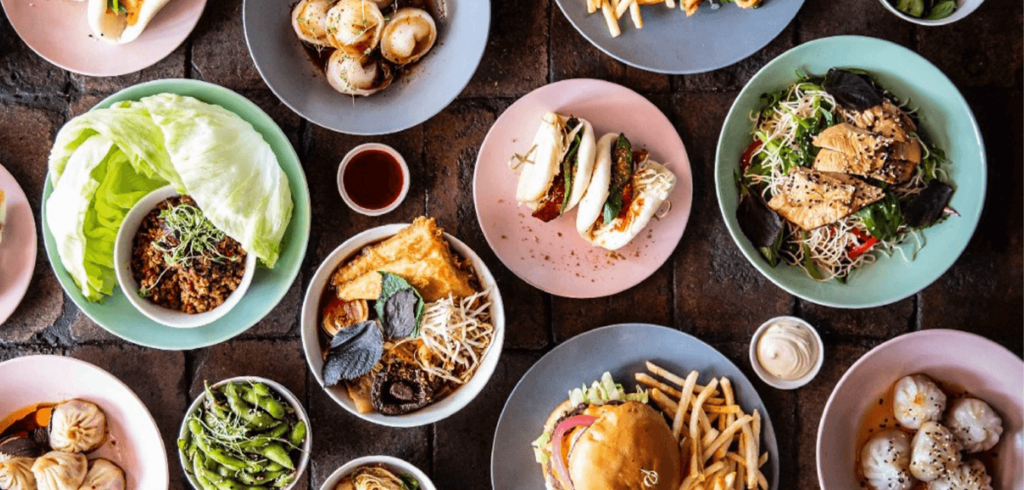 An overhead view of a variety of colorful plates filled with different foods, including dumplings, a burger with fries, salad, sandwiches, edamame, and seafood dishes, arranged on a dark tabletop.