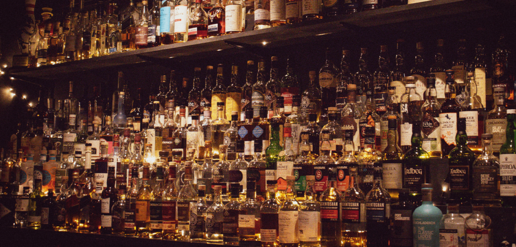 A dimly lit bar shelf filled with a variety of liquor bottles. The bottles are arranged in dense rows, showcasing different brands and types of alcohol, from whiskey to vodka. Warm lighting highlights the golden tones of the bottles.