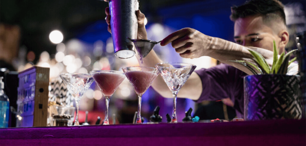 A bartender skillfully pouring cocktails through a strainer into martini glasses on a bar counter. The setting is dimly lit with vibrant purple and blue lighting, creating a lively atmosphere. A potted plant and other bar items are visible in the foreground.