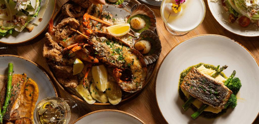 A table set with various seafood dishes, including lobster, scallops, and fish with asparagus, accompanied by a cocktail and a glass of white wine. Plates are artistically arranged on a wooden table.