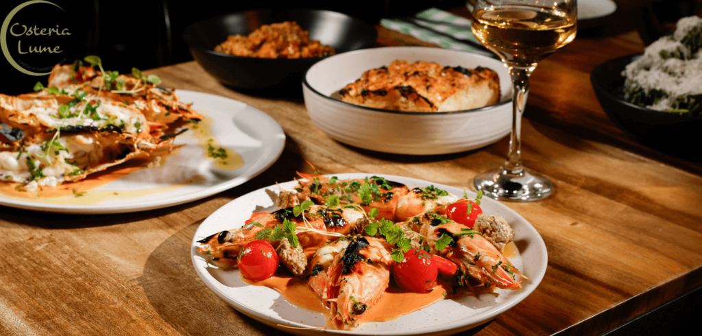 A table set with plates of grilled prawns garnished with herbs and cherry tomatoes, a dish of pasta, and a loaf of bread. A glass of white wine accompanies the meal. The Osteria Lume logo is seen in the upper left corner.