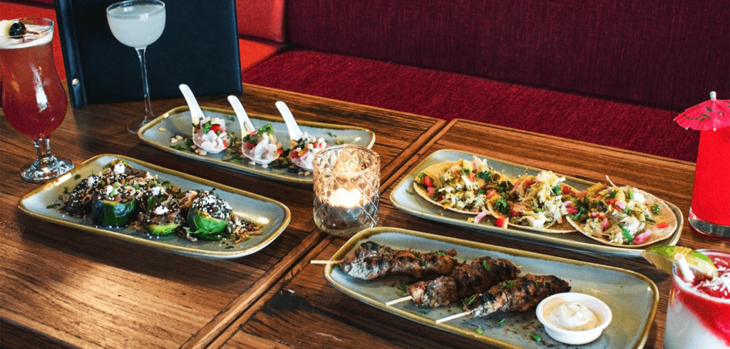 A wooden table is set with various dishes: three plates of appetizers, including skewers and tacos, alongside three different cocktails in glasses. A small lit candle adds ambiance to the scene.