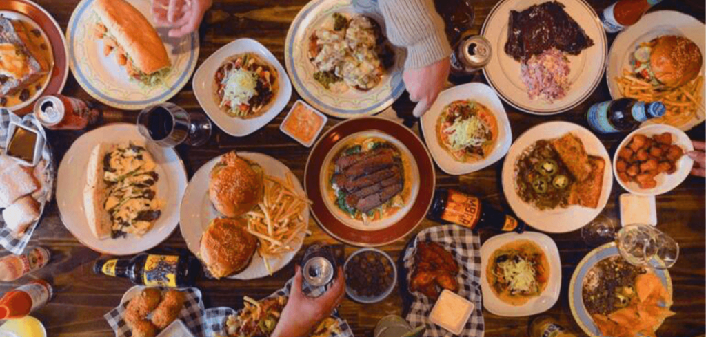 Aerial view of a table filled with various dishes, including burgers, fries, salads, sandwiches, steak, and beverages. Plates and glasses are scattered across the wooden surface, surrounded by people's hands reaching for food.